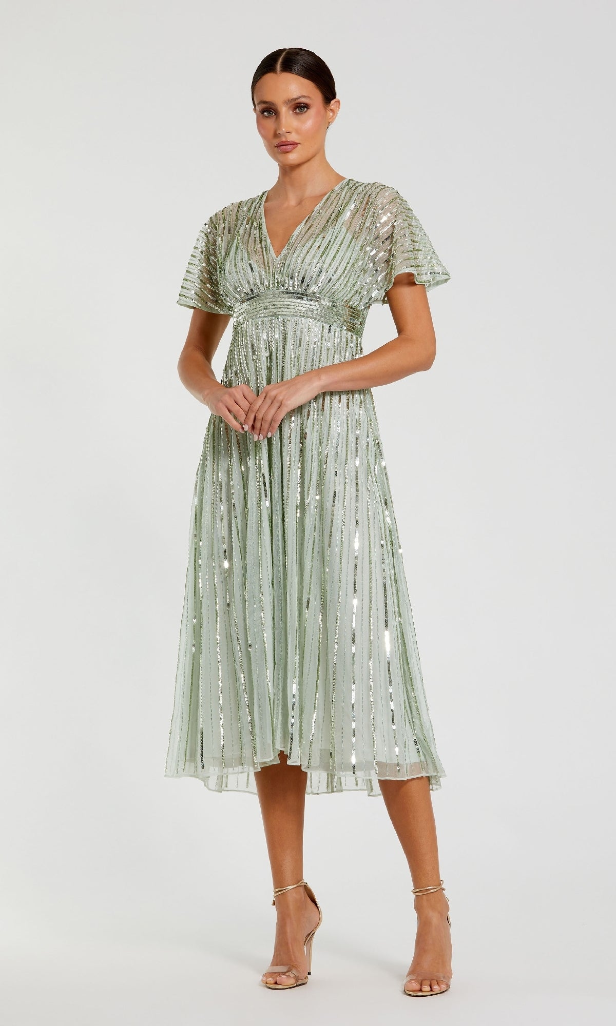 Woman wearing the Mac Duggal 14007 short sequin wedding-guest dress with light green V-neck, short sleeves, and sheer overlay, styled with strappy nude heels against a plain white background.